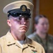 A U.S. Navy Chief Petty Officer in Service Khaki uniform stands at attention holding a folded American flag, suggesting a formal military ceremony such as a retirement or memorial service. The sailor displays multiple service ribbons, an aviation warfare device, and a surface warfare device on his uniform. A second uniformed individual is visible but out of focus in the background.