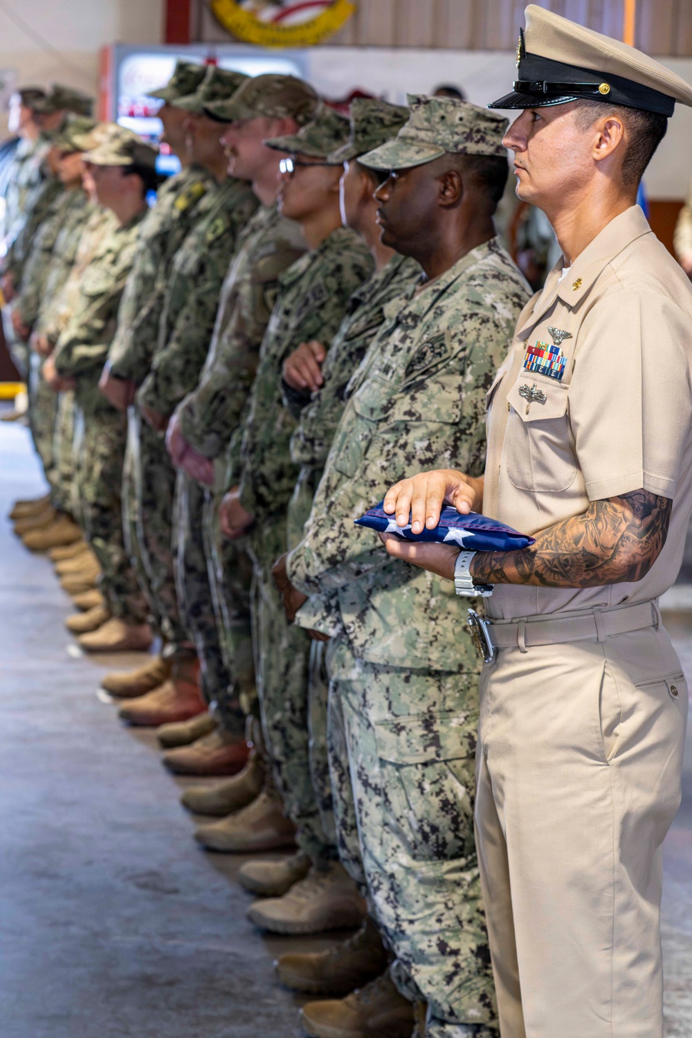 A formal military ceremony showing a line of service members standing at attention inside what appears to be a military facility. The foreground figure, wearing U.S. Navy Service Khaki uniform with a chief petty officer cover and multiple ribbons/badges, holds a folded American flag. Behind them, multiple personnel in Navy Working Uniform (NWU Type III) camouflage are lined up in formation.