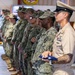 A formal military ceremony showing a line of service members standing at attention inside what appears to be a military facility. The foreground figure, wearing U.S. Navy Service Khaki uniform with a chief petty officer cover and multiple ribbons/badges, holds a folded American flag. Behind them, multiple personnel in Navy Working Uniform (NWU Type III) camouflage are lined up in formation.