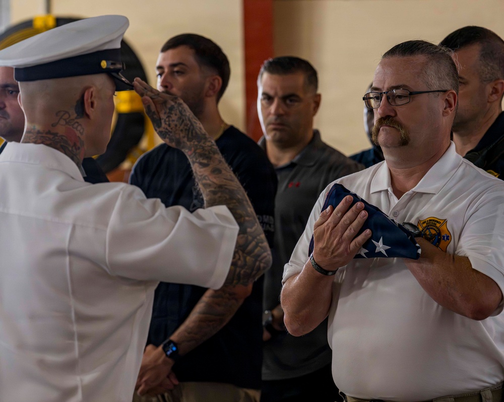 A formal military or first responder ceremony is taking place indoors, likely in a fire station or similar facility. A heavily tattooed individual in a white naval/dress uniform is rendering a salute while presenting a folded American flag to a man wearing a white polo shirt with what appears to be a fire/EMS department patch. Several other individuals in uniform and civilian attire stand solemnly in the background as witnesses.