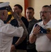A formal military or first responder ceremony is taking place indoors, likely in a fire station or similar facility. A heavily tattooed individual in a white naval/dress uniform is rendering a salute while presenting a folded American flag to a man wearing a white polo shirt with what appears to be a fire/EMS department patch. Several other individuals in uniform and civilian attire stand solemnly in the background as witnesses.