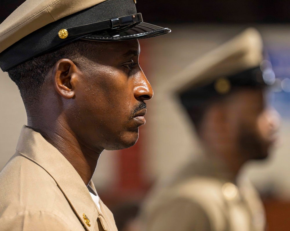 A close-up profile shot of a military service member in dress uniform, standing at attention. The individual is wearing a tan/khaki uniform with gold insignia and a peaked cap with a black brim and gold emblem. A second uniformed individual is visible but out of focus in the background.