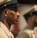 A close-up profile shot of a military service member in dress uniform, standing at attention. The individual is wearing a tan/khaki uniform with gold insignia and a peaked cap with a black brim and gold emblem. A second uniformed individual is visible but out of focus in the background.