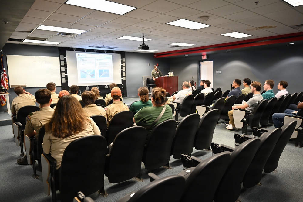 A presenter in military uniform stands at a wooden podium delivering a briefing or lecture to a mixed audience in a conference or briefing room. The audience includes individuals wearing Boy Scout uniforms as well as civilians seated in rows of black chairs. A projected slideshow with images and text is displayed on a screen at the front of the room.