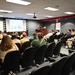 A presenter in military uniform stands at a wooden podium delivering a briefing or lecture to a mixed audience in a conference or briefing room. The audience includes individuals wearing Boy Scout uniforms as well as civilians seated in rows of black chairs. A projected slideshow with images and text is displayed on a screen at the front of the room.