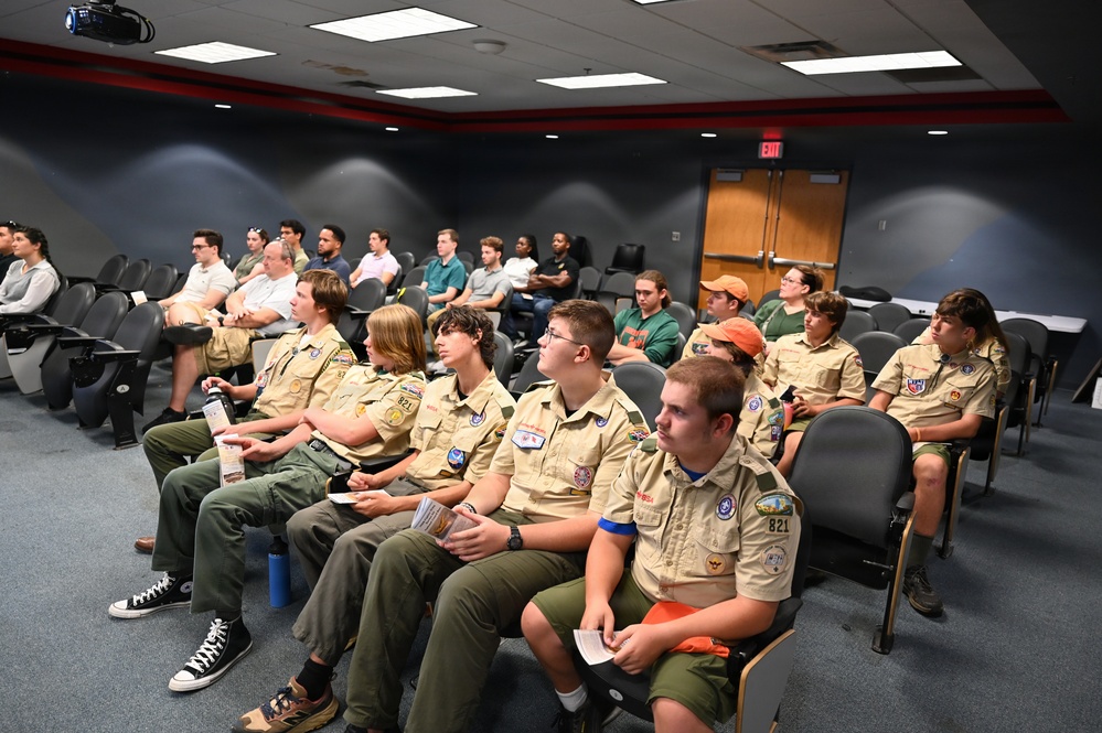 A group of Boy Scouts in tan uniforms with merit badge patches are seated in an auditorium or briefing room, attending a presentation or lecture. They are joined by other attendees in civilian clothing seated in rows behind them. The setting appears to be an official or institutional venue, possibly a government or military facility.