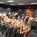 A group of Boy Scouts in tan uniforms with merit badge patches are seated in an auditorium or briefing room, attending a presentation or lecture. They are joined by other attendees in civilian clothing seated in rows behind them. The setting appears to be an official or institutional venue, possibly a government or military facility.