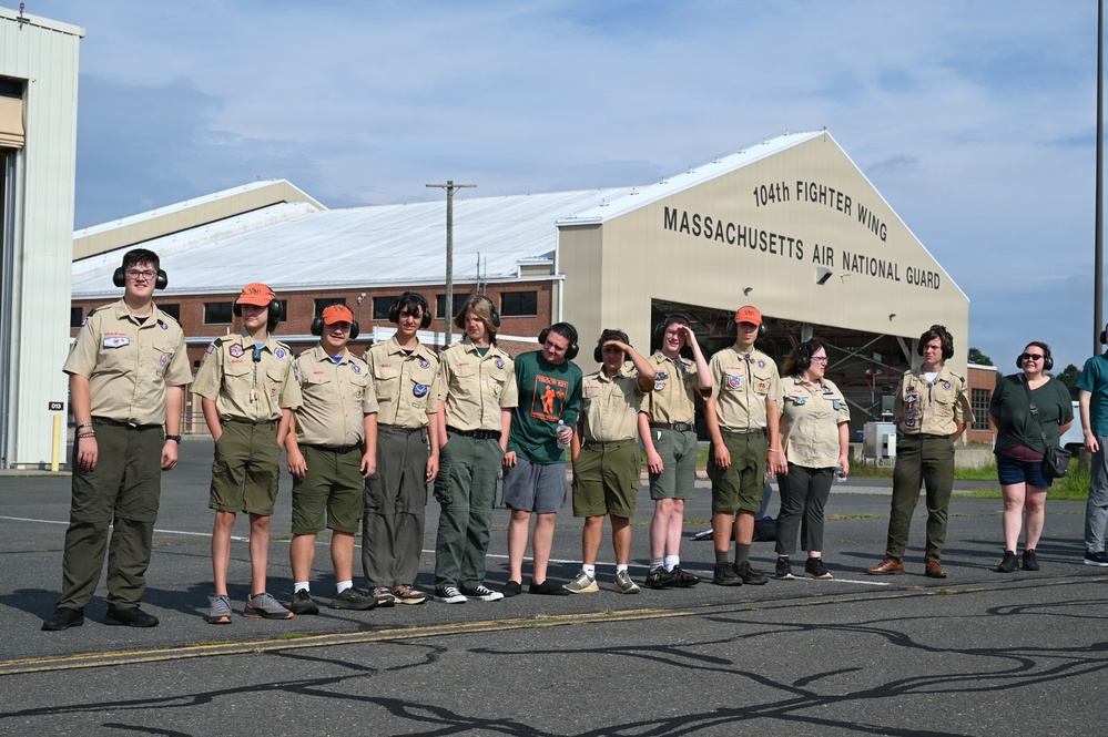 A group of Boy Scouts and adult leaders are standing on a tarmac in front of the 104th Fighter Wing Massachusetts Air National Guard hangar. Most participants are wearing tan Scout uniforms with olive green pants and orange caps, and nearly all are wearing hearing protection ear muffs. The group appears to be on an organized visit or tour of the military air base.