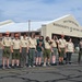 A group of Boy Scouts and adult leaders are standing on a tarmac in front of the 104th Fighter Wing Massachusetts Air National Guard hangar. Most participants are wearing tan Scout uniforms with olive green pants and orange caps, and nearly all are wearing hearing protection ear muffs. The group appears to be on an organized visit or tour of the military air base.
