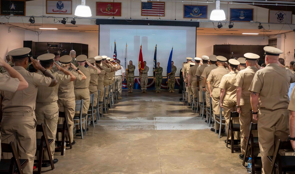 A military ceremony is taking place in an indoor hall with personnel in tan/khaki uniforms standing in two rows facing a central area where color guards hold flags. The projected screen at the front displays an image of the Twin Towers (World Trade Center), indicating this is a 9/11 memorial or remembrance ceremony. Personnel on both sides are rendering a salute toward the front of the room.