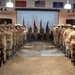A military ceremony is taking place in an indoor hall with personnel in tan/khaki uniforms standing in two rows facing a central area where color guards hold flags. The projected screen at the front displays an image of the Twin Towers (World Trade Center), indicating this is a 9/11 memorial or remembrance ceremony. Personnel on both sides are rendering a salute toward the front of the room.