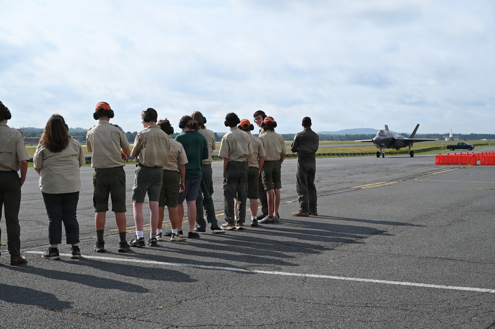 A group of youth scouts and adults stand on an airfield tarmac observing a military jet aircraft, likely an F-35, taxiing in the background. Several individuals are wearing orange hearing protection earmuffs, and a person in a flight suit stands to the right of the group. The scene appears to be an organized educational or familiarization visit to a military air base.