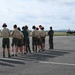 A group of youth scouts and adults stand on an airfield tarmac observing a military jet aircraft, likely an F-35, taxiing in the background. Several individuals are wearing orange hearing protection earmuffs, and a person in a flight suit stands to the right of the group. The scene appears to be an organized educational or familiarization visit to a military air base.