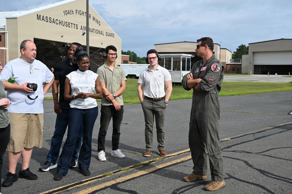 A military pilot in an olive green flight suit is briefing a group of civilians on the tarmac of the 104th Fighter Wing, Massachusetts Air National Guard base. The group of approximately six civilians appears to be on an official visit or tour of the facility. The setting is outdoors near aircraft hangars on a partly cloudy day.