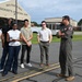 A military pilot in an olive green flight suit is briefing a group of civilians on the tarmac of the 104th Fighter Wing, Massachusetts Air National Guard base. The group of approximately six civilians appears to be on an official visit or tour of the facility. The setting is outdoors near aircraft hangars on a partly cloudy day.