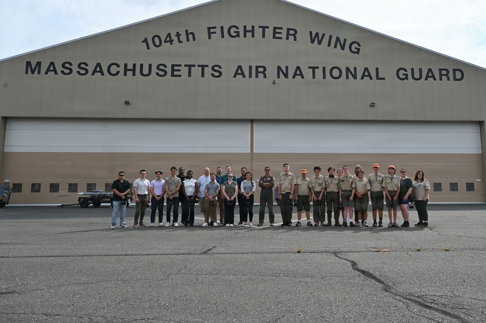 A group of approximately 25 people pose for a group photo in front of the 104th Fighter Wing, Massachusetts Air National Guard hangar. The group includes individuals in Boy Scout uniforms, civilian clothing, and at least one person in a flight suit. The setting appears to be a military airbase tarmac during a visit or tour.