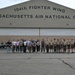 A group of approximately 25 people pose for a group photo in front of the 104th Fighter Wing, Massachusetts Air National Guard hangar. The group includes individuals in Boy Scout uniforms, civilian clothing, and at least one person in a flight suit. The setting appears to be a military airbase tarmac during a visit or tour.