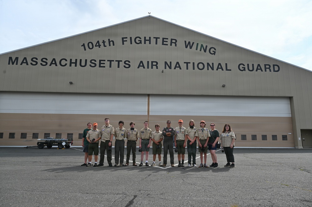 A group of approximately 13 individuals, mostly wearing Boy Scout uniforms, pose for a group photo in front of the 104th Fighter Wing Massachusetts Air National Guard hangar. One person in the center appears to be wearing a military flight suit, likely a guard member serving as a host or guide. The visit appears to be an organized Scout troop tour of the Air National Guard facility.