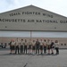 A group of approximately 13 individuals, mostly wearing Boy Scout uniforms, pose for a group photo in front of the 104th Fighter Wing Massachusetts Air National Guard hangar. One person in the center appears to be wearing a military flight suit, likely a guard member serving as a host or guide. The visit appears to be an organized Scout troop tour of the Air National Guard facility.
