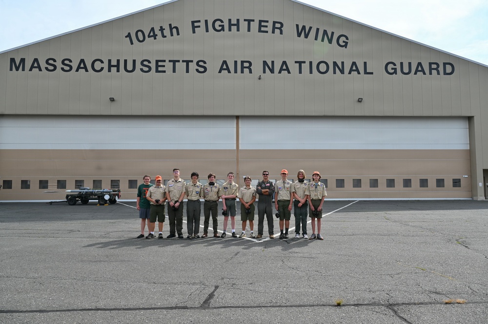 A group of approximately 11 individuals, mostly wearing Boy Scout uniforms, pose for a group photo in front of the 104th Fighter Wing Massachusetts Air National Guard hangar. One person in the center appears to be wearing a military flight suit, likely a National Guard pilot or personnel serving as a host. The setting is a military airfield tarmac with a large hangar door visible in the background.
