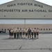 A group of approximately 11 individuals, mostly wearing Boy Scout uniforms, pose for a group photo in front of the 104th Fighter Wing Massachusetts Air National Guard hangar. One person in the center appears to be wearing a military flight suit, likely a National Guard pilot or personnel serving as a host. The setting is a military airfield tarmac with a large hangar door visible in the background.