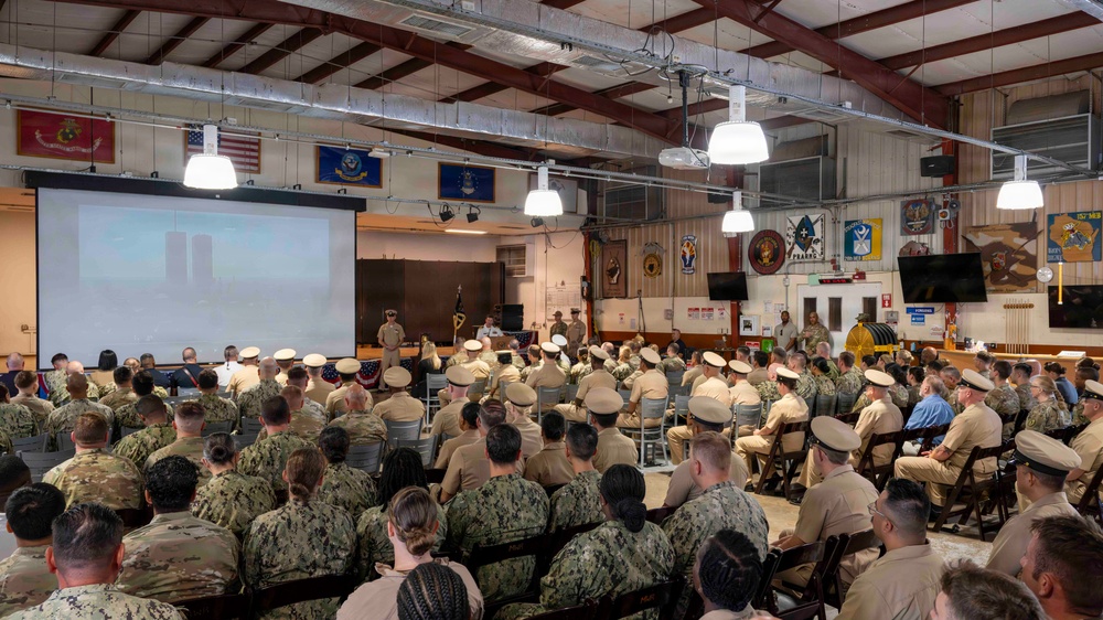A large group of military personnel are gathered in what appears to be a base assembly hall or community center for a formal briefing or ceremony. The audience consists of sailors in both Navy Working Uniform (NWU) digital camouflage and khaki service uniforms, seated in rows facing a projection screen and stage area. Multiple military branch flags and unit insignia are displayed on the walls throughout the facility.