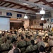 A large group of military personnel are gathered in what appears to be a base assembly hall or community center for a formal briefing or ceremony. The audience consists of sailors in both Navy Working Uniform (NWU) digital camouflage and khaki service uniforms, seated in rows facing a projection screen and stage area. Multiple military branch flags and unit insignia are displayed on the walls throughout the facility.