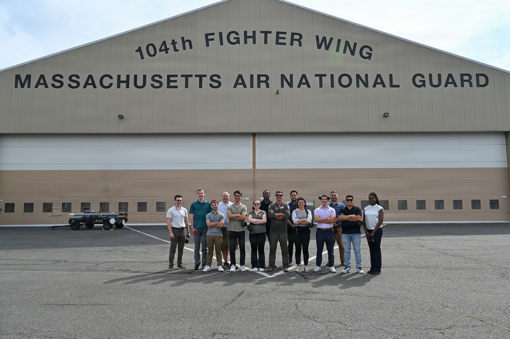 A group of approximately 14 individuals poses for a group photo in front of a large military hangar belonging to the 104th Fighter Wing, Massachusetts Air National Guard. The group appears to be a mix of civilians and at least one uniformed military personnel, standing on a tarmac apron. The setting suggests an official visit, tour, or outreach event at the air base.