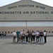 A group of approximately 14 individuals poses for a group photo in front of a large military hangar belonging to the 104th Fighter Wing, Massachusetts Air National Guard. The group appears to be a mix of civilians and at least one uniformed military personnel, standing on a tarmac apron. The setting suggests an official visit, tour, or outreach event at the air base.