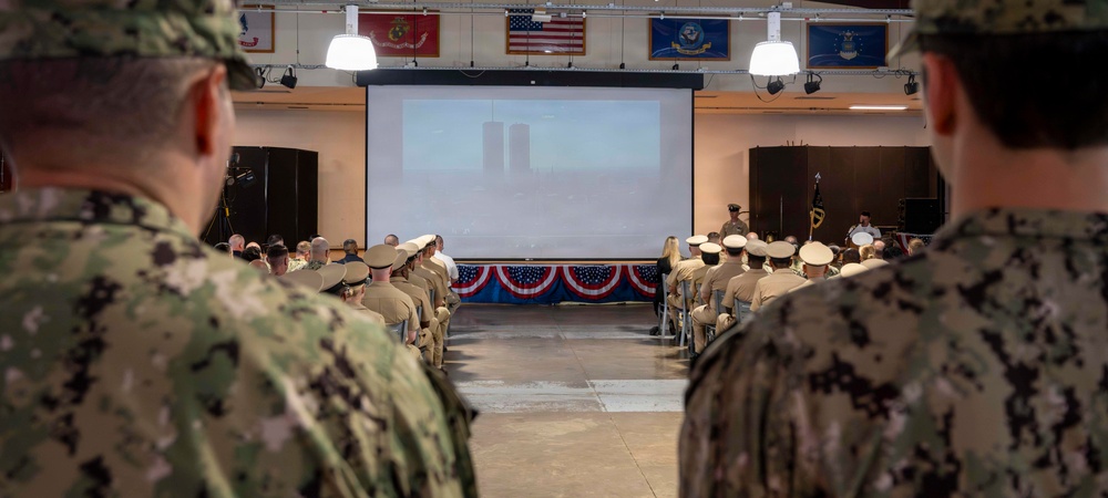 Military personnel in camouflage and dress uniforms are gathered in a ceremonial hall for what appears to be a memorial or commemorative event. A projection screen at the front displays an image of the New York City skyline featuring the Twin Towers, suggesting this is a 9/11 remembrance ceremony. The venue is decorated with American patriotic bunting and displays multiple military branch flags.