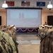 Military personnel in camouflage and dress uniforms are gathered in a ceremonial hall for what appears to be a memorial or commemorative event. A projection screen at the front displays an image of the New York City skyline featuring the Twin Towers, suggesting this is a 9/11 remembrance ceremony. The venue is decorated with American patriotic bunting and displays multiple military branch flags.