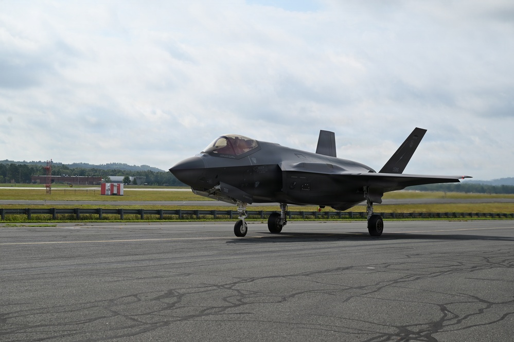 A dark-colored F-35 Lightning II stealth fighter jet is taxiing on an airport or airbase tarmac. The aircraft is photographed from a front-left angle, showing its distinctive stealth design with swept wings and angular surfaces. The setting appears to be a military or joint-use airfield with green terrain and overcast skies in the background.