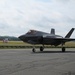 A dark-colored F-35 Lightning II stealth fighter jet is taxiing on an airport or airbase tarmac. The aircraft is photographed from a front-left angle, showing its distinctive stealth design with swept wings and angular surfaces. The setting appears to be a military or joint-use airfield with green terrain and overcast skies in the background.