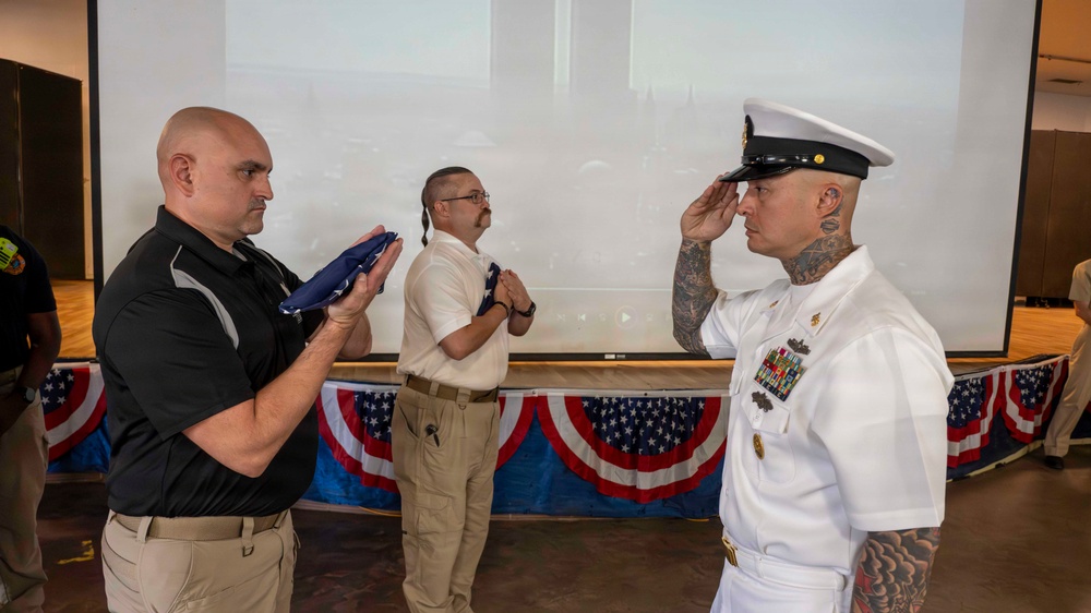 A formal military ceremony is taking place indoors, likely a retirement or award ceremony. A U.S. Navy senior enlisted sailor in dress whites is rendering a salute while a civilian holds a folded American flag. The setting is decorated with patriotic bunting along a stage, suggesting an official commemorative event.