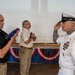 A formal military ceremony is taking place indoors, likely a retirement or award ceremony. A U.S. Navy senior enlisted sailor in dress whites is rendering a salute while a civilian holds a folded American flag. The setting is decorated with patriotic bunting along a stage, suggesting an official commemorative event.