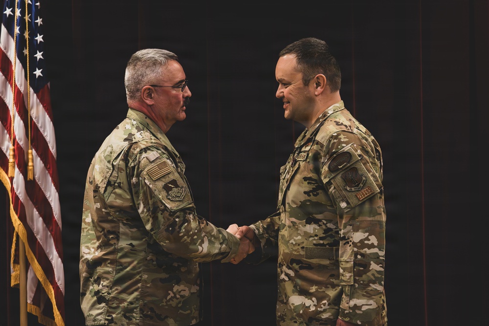 Two military personnel in OCP (Operational Camouflage Pattern) uniforms are shaking hands in what appears to be a formal military ceremony. Both individuals are wearing U.S. Air Force uniforms with patches visible on their sleeves. An American flag with gold fringe is prominently displayed in the background to the left.