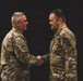 Two military personnel in OCP (Operational Camouflage Pattern) uniforms are shaking hands in what appears to be a formal military ceremony. Both individuals are wearing U.S. Air Force uniforms with patches visible on their sleeves. An American flag with gold fringe is prominently displayed in the background to the left.