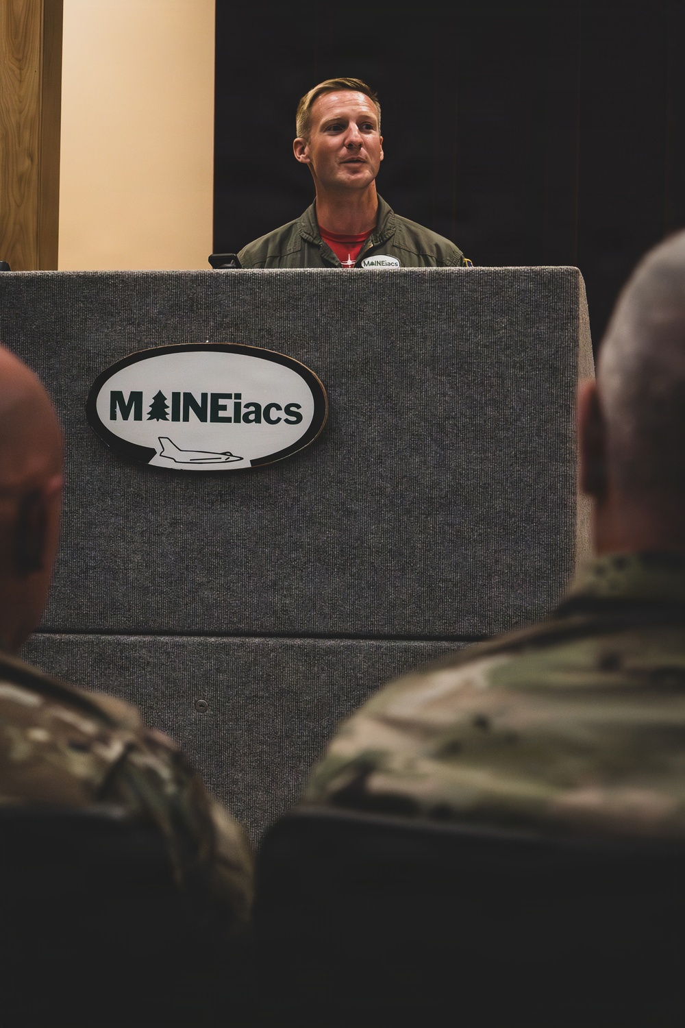 A military aviator in an olive green flight suit stands at a podium bearing the 'MAINEiacs' logo, which features a pine tree and a fighter jet silhouette. He appears to be addressing an audience that includes personnel in camouflage uniforms visible in the foreground. The setting appears to be a formal military briefing or presentation event.