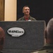 A military aviator in an olive green flight suit stands at a podium bearing the 'MAINEiacs' logo, which features a pine tree and a fighter jet silhouette. He appears to be addressing an audience that includes personnel in camouflage uniforms visible in the foreground. The setting appears to be a formal military briefing or presentation event.