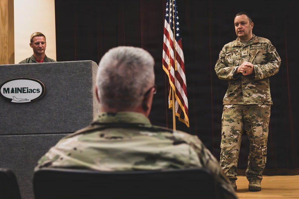 A military briefing or presentation is taking place in what appears to be an auditorium or meeting hall. A U.S. Air Force officer in OCP (Operational Camouflage Pattern) uniform stands near an American flag and addresses an audience, while another service member sits at a podium bearing the 'MAINEiacs' insignia. A third military member is visible seated in the foreground with their back to the camera.