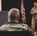 A military briefing or presentation is taking place in what appears to be an auditorium or meeting hall. A U.S. Air Force officer in OCP (Operational Camouflage Pattern) uniform stands near an American flag and addresses an audience, while another service member sits at a podium bearing the 'MAINEiacs' insignia. A third military member is visible seated in the foreground with their back to the camera.