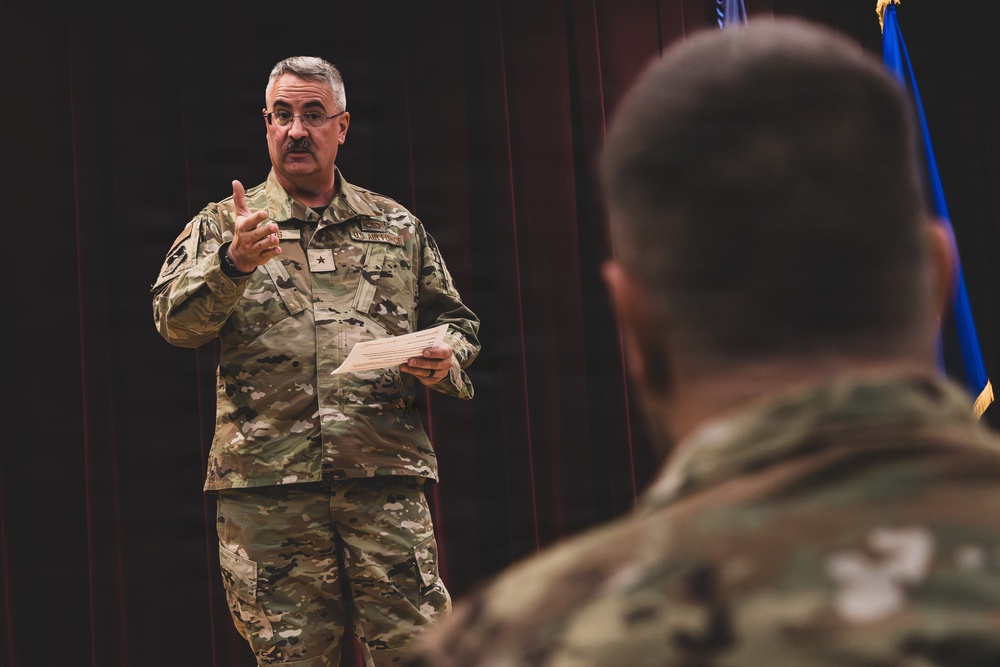 A U.S. military officer in OCP (Operational Camouflage Pattern) uniform is addressing an audience, holding papers and gesturing with one hand. The speaker appears to be a General officer based on the visible star insignia on the chest. The setting appears to be an auditorium or formal briefing room with a dark curtain backdrop and a blue flag visible in the background.