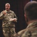 A U.S. military officer in OCP (Operational Camouflage Pattern) uniform is addressing an audience, holding papers and gesturing with one hand. The speaker appears to be a General officer based on the visible star insignia on the chest. The setting appears to be an auditorium or formal briefing room with a dark curtain backdrop and a blue flag visible in the background.