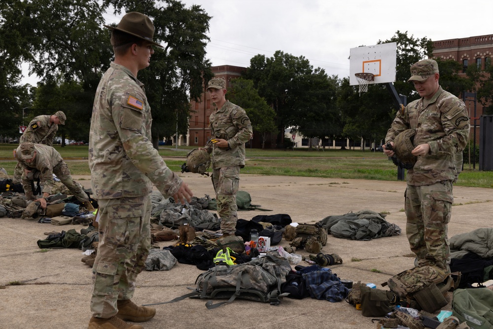 A group of U.S. Army soldiers in MultiCam/OCP camouflage uniforms are gathered on an outdoor paved area, appearing to conduct a gear inspection or layout drill. One soldier wearing a drill sergeant hat is gesturing and appears to be giving instructions to others standing near piles of equipment and backpacks spread on the ground. The setting appears to be a military installation or ROTC training area, with a basketball hoop and brick buildings visible in the background.