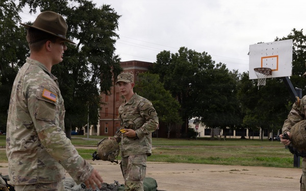 A group of U.S. Army soldiers in MultiCam/OCP camouflage uniforms are gathered on an outdoor paved area, appearing to conduct a gear inspection or layout drill. One soldier wearing a drill sergeant hat is gesturing and appears to be giving instructions to others standing near piles of equipment and backpacks spread on the ground. The setting appears to be a military installation or ROTC training area, with a basketball hoop and brick buildings visible in the background.
