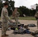 A group of U.S. Army soldiers in MultiCam/OCP camouflage uniforms are gathered on an outdoor paved area, appearing to conduct a gear inspection or layout drill. One soldier wearing a drill sergeant hat is gesturing and appears to be giving instructions to others standing near piles of equipment and backpacks spread on the ground. The setting appears to be a military installation or ROTC training area, with a basketball hoop and brick buildings visible in the background.