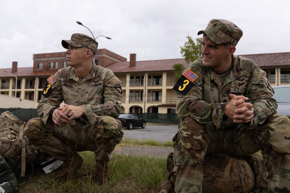 Two U.S. Army soldiers in Operational Camouflage Pattern (OCP) uniforms are crouching outdoors on a military installation. Both wear matching unit patches including a black '3' numeral patch and U.S. flag patches on their left shoulders. They appear to be resting or observing something off-camera in front of a large military barracks building.