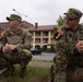 Two U.S. Army soldiers in Operational Camouflage Pattern (OCP) uniforms are crouching outdoors on a military installation. Both wear matching unit patches including a black '3' numeral patch and U.S. flag patches on their left shoulders. They appear to be resting or observing something off-camera in front of a large military barracks building.