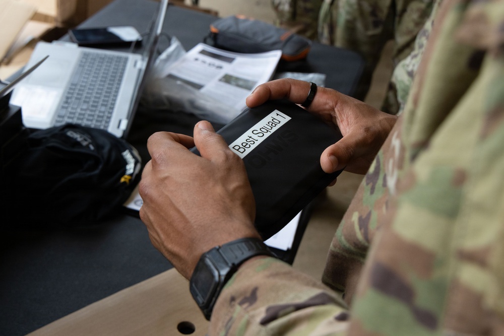 A soldier in MultiCam/OCP camouflage uniform is holding a black device or case labeled 'Best Squad 1' with a printed adhesive label. The setting appears to be an administrative or operations table with a laptop, documents, and equipment visible in the background. The image appears to document a military training or competition event.