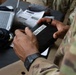 A soldier in MultiCam/OCP camouflage uniform is holding a black device or case labeled 'Best Squad 1' with a printed adhesive label. The setting appears to be an administrative or operations table with a laptop, documents, and equipment visible in the background. The image appears to document a military training or competition event.